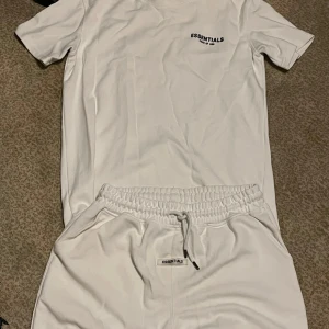 Vit Essentials Fear of God Set - Säljer en clean vit Ser från Essentials Fear of God. Settet har svart logga tryckt på bröstet och ryggen, klassisk rund hals och korta ärmar. Skön passform och tillverkad i mjuk bomull, perfekt för en chill och stilren look.