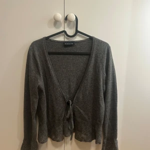 Grå kofta med knyt från Birgitta - Mysig grå kofta från Birgitta i ullmix med v-ringning och knyt framtill. Fina virkade detaljer vid ärmslut och nederkant ger en extra touch. Perfekt för lager-på-lager och chill höst- eller vinterstil.