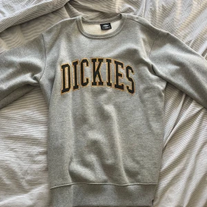  sweatshirt Dickies  - Helt oanvänd Snygg grå sweatshirt från Dickies i storlek L. Tröjan har klassisk rund halsringning och stort broderat Dickies-tryck i svart och gult på bröstet. Mjuk insida och ribbade muddar vid ärmslut och nederkant. 