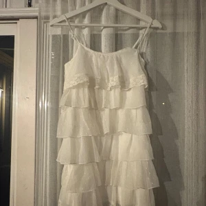 Vit volangklänning från Lindex - Supersöt vit kortklänning från Lindex med tunna axelband och flera lager volanger. Klänningen har dragkedja i ryggen och är tillverkad i ett lätt, flowy material som känns luftigt. Perfekt för sommarens alla tillfällen när du vill känna dig extra fin. En liten fläck under ena volangen