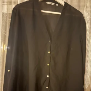 Svart skjorta med knappar - Stilren svart skjorta från Vero Moda med v-ringning och knappar framtill. Skjortan har lång ärm och en klassisk passform, perfekt för dig som gillar en clean och enkel look. Materialet känns lätt och luftigt.