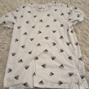 Vit Adidas t-shirt med logomönster - Snygg vit t-shirt från Adidas med svart logomönster över hela plagget. Klassisk rund halsringning och korta ärmar. Tillverkad i mjuk bomull som är skön mot huden. Perfekt för en sportig och avslappnad stil.