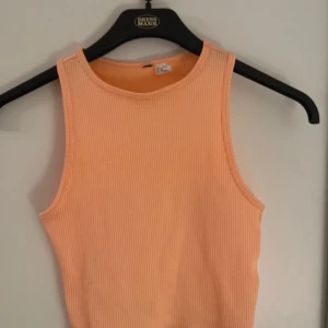 Ribbad persikofärgad linnetopp - Säljer en ribbad linnetopp i en snygg persikoorange färg. Toppen är ärmlös med rund halsringning och croppad passform, perfekt för varma dagar. Materialet är stretchigt och bekvämt, vilket gör den enkel att styla med jeans eller kjol.