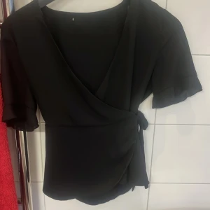 Svart omlottblus med knytband - Snygg svart omlottblus med korta ärmar och volangkant. Blusen har knytband i midjan och en v-ringad omlottfront som ger en stilren look. Perfekt att styla med jeans eller kjol för en trendig outfit.