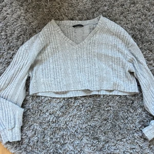 Grå croppad stickad tröja Shein - Säljer en ljusgrå croppad stickad tröja från Shein med v-ringning och ribbat mönster. Tröjan har långa ärmar och en loose passform, perfekt för lager-på-lager och chill dagar.