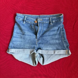 Högmidjade jeansshorts från H&M Divided - Snygga blå jeansshorts från H&M Divided i storlek 36. De är högmidjade med klassisk femficksdesign, uppvikta benslut och stängs med knapp och dragkedja. Perfekta för varma dagar och enkel att matcha med allt.