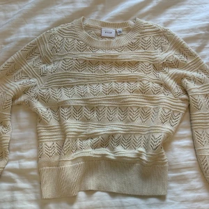 Beige stickad tröja från Vila - Superfin beige stickad tröja från Vila med snyggt hålmönster och rund halsringning. Tröjan har ribbade muddar och ett luftigt, mönstrat stickat tyg som ger en fräsch och trendig look. Perfekt att styla med jeans eller kjol.