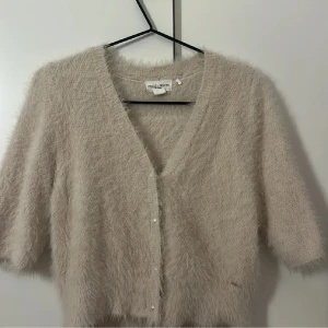 Beige fluffig kofta  - Supermjuk och fluffig beige kofta från House of White med v-ringning och pärlknappar. Koftan har kortare ärmar och en croppad passform som ger en trendig look. Perfekt att slänga över en topp för extra mysfaktor.