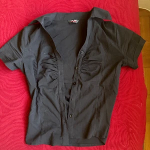 Svart kortärmad skjorta från ÜZ-SA - Svart skjorta från ÜZ-SA med korta ärmar och klassisk krage. Skjortan har knappar framtill och snygga rynkade detaljer vid bysten som ger en cool och modern look. Perfekt för dig som gillar stilrena plagg med en twist. Ska sitta lite mer tight. Saknar en knapp men knappt använd. Känns som en storlek M
