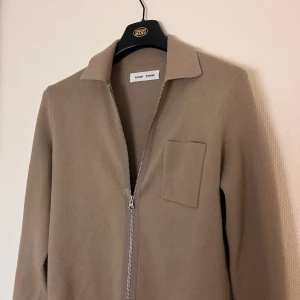 Beige overshirt från Samsøe Samsøe - Snygg beige overshirt från Samsøe Samsøe med klassisk krage, bröstficka och hel dragkedja framtill. Jackan har en stilren och minimalistisk look som passar perfekt till lager-på-lager. Materialet är mjukt och har en exklusiv känsla.