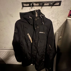 Svart Didriksons parkajacka, strl 38 - Svart parkajacka från Didriksons med huva, dragkedja och tryckknappar. Jackan har flera fickor, stormsystem-foder och snygga detaljer i beige. Perfekt för kalla dagar och har en klassisk, stilren look.