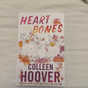 Heart Bones - En gripande roman av Colleen Hoover, perfekt för unga vuxna. Omslaget pryds av färgstarka blommor och två dödskallar, vilket ger en känsla av både skörhet och styrka. En bok som lockar till läsning om känslor och relationer.