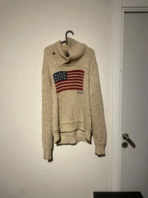 Beige polotröja Polo Ralph Lauren S - Stickad beige polotröja från Polo Ralph Lauren i storlek S. Tröjan har en bred, ribbad polokrage med knappdetalj och ett stort amerikanskt flaggmotiv i blått, vitt och rött framtill. Mjukt material och ribbade muddar vid ärmslut och nederkant.