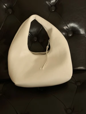 Vit axelväska i skinn - Snygg vit axelväska med mjuk, rundad form och minimalistisk design. Väskan är tillverkad i slätt skinn och har en praktisk dragkedja upptill med dekorativt snöre. Perfekt storlek för att få plats med allt du behöver.