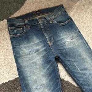 Nudie Jeans Blå 28/32 high Kai - Snygga blåa jeans från Nudie Jeans med slitna detaljer och klassisk femficksdesign. Jeansen har orangea sömmar, normalhög midja och är tillverkade i bomull. Perfekta för dig som gillar en smal passform och cool tvätt. Säljer dessa för de inte passar, model high Kai.