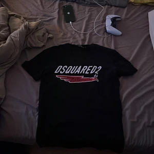 Svart DSQUARED2 t-shirt med print - Svart t-shirt från DSQUARED2 i storlek M med vit logotyp och röd vinge framtill. Klassisk rund hals och korta ärmar. Snygg och stilren design som sticker ut med det grafiska trycket. Tillverkad i mjuk bomull för skön känsla.