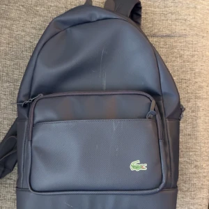 Svart ryggsäck från Lacoste - Snygg svart ryggsäck från Lacoste med klassisk krokodillogga på framsidan. Väskan har ett stort huvudfack, en praktisk ytterficka med dragkedja och justerbara axelremmar. Materialet är slitstark polyester och insidan har en diskret Lacoste-label.