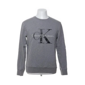 Grå sweatshirt från Calvin Klein Jeans - Snygg grå sweatshirt från Calvin Klein Jeans med stort CK-tryck på bröstet. Tröjan har rund hals, långa ärmar och ribbade muddar vid ärmslut och nederkant. Tillverkad i 100% bomull för skön komfort och stilren look.