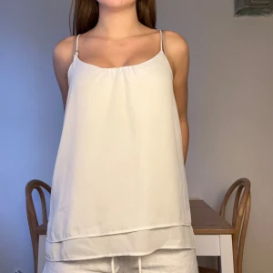 Vit linnetopp med tunna axelband - Säljer en stilren vit linnetopp med tunna justerbara axelband och lätt rynkning vid halsringningen. Toppen har en luftig passform och dubbla lager tyg nedtill för extra detalj. Perfekt för dig som gillar enkel och fräsch stil.