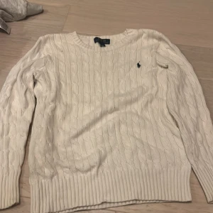 Vit kabelstickad tröja Polo Ralph Lauren - Säljer en vit kabelstickad tröja från Polo Ralph Lauren i storlek 10-12 år.Tröjan har rund hals, ribbade muddar och det klassiska Polo-logot på bröstet. Perfekt för dig som gillar stilrena och tidlösa plagg med snygg struktur.