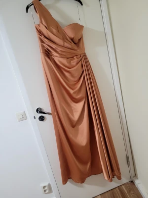 Champagnefärgad långklänning oneshoulder - Säljer en elegant champagnefärgad långklänning med oneshoulder-design och draperade detaljer över byst och midja. Klänningen har en snygg omlott-effekt och ett mjukt, glansigt tyg som faller fint. Perfekt för dig som vill sticka ut med stil på bal eller fest.