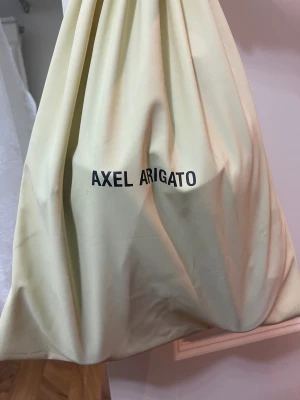 Svarta sneakers Axel Arigato mocka - Stilrena svarta sneakers från Axel Arigato i mjuk mocka med diskret guldfärgad logga på sidan. Skorna har vit platt sula och minimalistisk design utan synlig snörning. Kommer med original dustbag. Perfekta för dig som gillar clean och modern look. Passar ganska bra med kostym och liknande stiliga kläder 