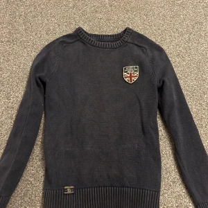 Mörkblå stickad tröja från Morris - Snygg mörkblå stickad tröja från Morris Gents i storlek XS. Tröjan har ett broderat märke med brittisk flagga på bröstet och ribbade muddar vid hals, ärmslut och nederkant. Perfekt för dig som gillar klassisk och stilren look.