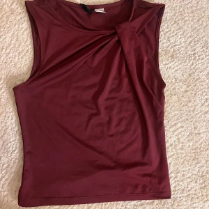 Vinröd ärmlös topp med twist - Snygg vinröd topp med twistad detalj vid axeln. Modellen är ärmlös och har en rund halsringning. Materialet känns mjukt och stretchigt, perfekt för att skapa en cool och stilren look. Aldrig andvänt! Bara att skriva för flera bilder eller funderingar🫰🏼🫰🏼