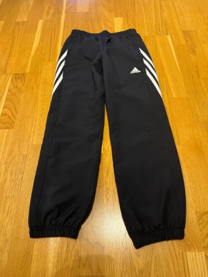 Svarta Adidas träningsbyxor/vindbyxor strl 152 barn - Svarta träningsbyxor/vindbyxor från Adidas med klassiska vita ränder längs sidorna och broderad logga. Byxorna har elastisk midja, dragkedjor vid fickorna.