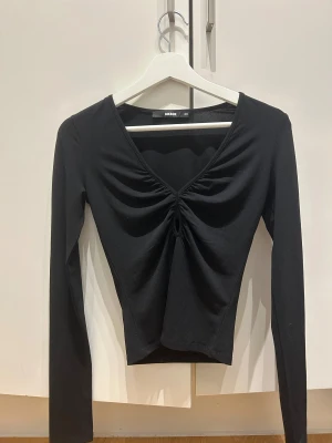 Svart långärmad topp från Bik Bok - Snygg svart långärmad topp från Bik Bok med v-ringning och cool snörningsdetalj framtill. Modellen är croppad och har en slim passform som framhäver silhuetten. Perfekt för dig som gillar stilrena och trendiga plagg.