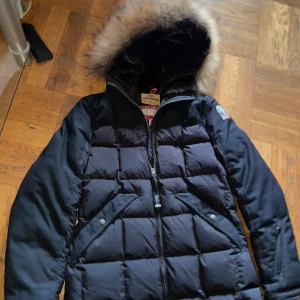 Svart Parajumpers dunjacka med päls  - Snygg svart dunjacka från Parajumpers med quiltad design och fluffig päls runt huvan. Jackan har dragkedja framtill, två fickor med tryckknappar och ett broderat Parajumpers-märke på ärmen. Perfekt för kalla dagar när du vill hålla stilen.