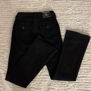 Svarta True Religion bootcut jeans - Snygga svarta jeans från True Religion, modell Becca Mid Rise Bootcut. Klassisk femficksdesign med unika bakfickor med lock och knapp. Jeansen har medelhög midja och är tillverkade i ett mjukt bomullsmaterial med stretch för skön passform. Jag är 164cm och byxan är en aning lång på mig, därav är den lite missfärgade längst ner. Säljer dessa byxor då dem inte kommer till så mycket användning för att jag tycker de är för hög midja på dem. Byxorna passar någon som gillar stilrena och mjuka jeans. 