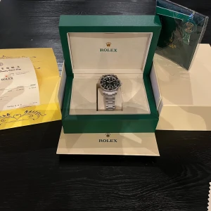 Rolex Submariner svart/silver klocka - Rolex Submariner med svart urtavla och silverfärgad länk i rostfritt stål. Klassisk rund form, tydliga index och datumfönster. Snygg och ikonisk design med vridbar bezel och robust spänne. Levereras med originalbox och certifikat.