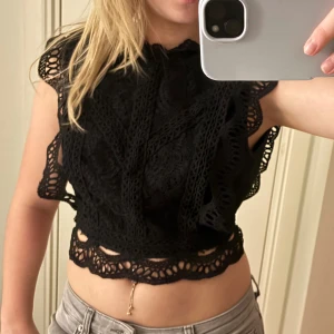 Svart spetsig croppad blus - Säljer en svart croppad blus med spetsdetaljer och volangkant. Toppen har ärmlös design med snygga virkade partier och öppen rygg med knappstängning. Perfekt för dig som gillar unika detaljer och vill sticka ut.