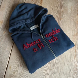 Abercrombie & Fitch zip up - Mörkblå hoodie från Abercrombie & Fitch med röd broderad logga på bröstet och en liten röd älg på ärmen. Tröjan har huva med snörning, dragkedja framtill och är fodrad med mjuk fleece. Perfekt för chill dagar och snygg streetstil. 60 bredd 70 längd