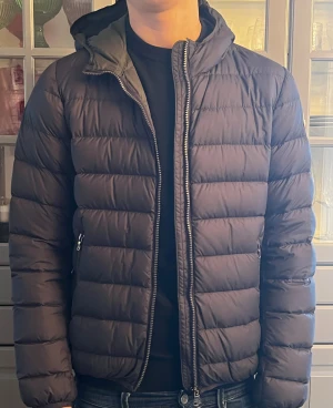 Mörkblå Colmar dunjacka med huva - Snygg mörkblå dunjacka från Colmar i storlek 16y, men är som en storlek Small. Jackan har en vadderad, quiltad design med huva, dragkedja framtill och två sidofickor med zip. Perfekt för kalla dagar och har en clean look med Colmar-logga på ärmen.
