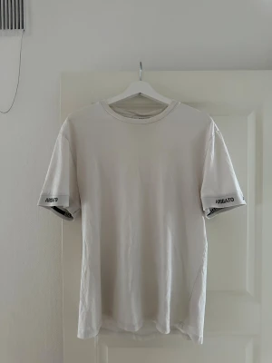 Vit t-shirt från Axel Arigato - Snygg vit t-shirt från Axel Arigato med tryckt logga på ärmsluten. Klassisk rund hals och rak passform. Materialet är mjuk bomull som känns skönt mot huden. Perfekt för dig som gillar stilrena och cleana plagg med diskreta detaljer.