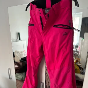 Rosa täckbyxor från McKinley, stl 152 - Säljer ett par knallrosa täckbyxor från McKinley i storlek 152. Byxorna har svarta detaljer, dragkedjor vid fickorna och justerbara hällor i midjan. Perfekta för vintern och skidbacken, med slitstarkt syntetmaterial som håller dig varm och torr.
