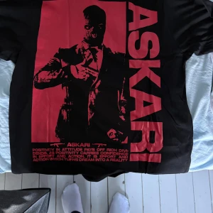 Svart t-shirt med röd print från Askari - Cool svart t-shirt från Askari med stor röd print på framsidan som visar en person i kostym och texten ASKARI. På bröstet finns en mindre röd logga med texten 'ASKARI ONE MAN ARMY' och ett vapenmotiv. Gjord i 100% ekologisk bomull, rund hals och korta ärmar.