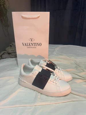 Valentino Garavani Open sneakers vit/blå - Snygga Valentino Garavani Open sneakers i vitt skinn med bred marinblå rem över mitten. Klassisk rund tå, platt sula och vita snören. Lyxig känsla med guldfärgad logga på innersulan. Perfekt för dig som gillar stilrena och exklusiva sneakers. Passar någon med 42-43 i storlek, endast testade utomhus 1 gång då dem var för små för mig. Låda finns inte.