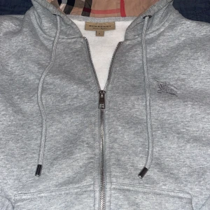 Grå Burberry hoodie med dragkedja - Snygg grå hoodie från Burberry med klassiskt rutigt foder i huvan och broderad logga på bröstet. Tröjan har dragkedja framtill, snörning vid huvan och är tillverkad i mjuk bomull. Perfekt för dig som gillar stilrena detaljer och exklusiva märken.