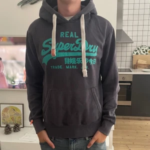 Mörkgrå Superdry hoodie med turkos tryck - Säljer en mörkgrå hoodie från Superdry med turkost tryck och japanska tecken på bröstet. Tröjan har vit snörning i huvan, känguruficka och ribbade muddar. Perfekt för dig som gillar streetwear och vill ha en snygg och bekväm look. Modellen är 185. 