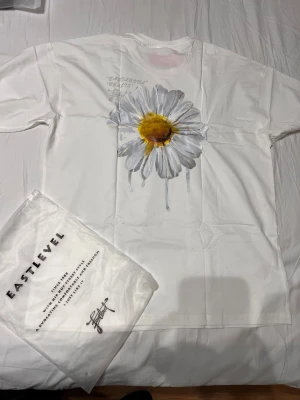 Vit t-shirt med prästkrage från Eastlevel - Vit t-shirt från Eastlevel med stor tryckt prästkrage i grått och gult på framsidan. Klassisk passform med korta ärmar och rund hals. Materialet är mjuk bomull, perfekt för en chill och stilren look.