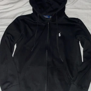 Svart hoodie från Polo Ralph Lauren - Svart hoodie från Polo Ralph Lauren med vit broderad logga på bröstet. Tröjan har huva med snörning, dragkedja framtill och två fickor. Materialet är mjuk bomull och passformen är normal. Perfekt för chill dagar och streetwear.
