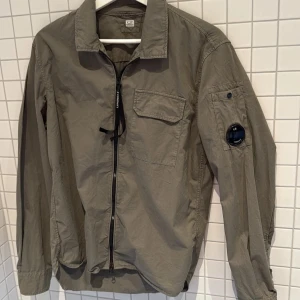 C.P. Company overshirt olivgrön -  Snygg olivgrön overshirt från C.P. Company med klassisk krage, bröstficka och ikonisk linsdetalj på ärmen. Jackan har dragkedja framtill, perfekt för lager-på-lager och en clean streetstyle-look.