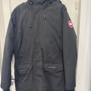 Svart parkajacka från Canada Goose XL - Svart parkajacka från Canada Goose i storlek L. Jackan har huva med snörning, flera fickor och klassisk Canada Goose patch på ärmen. Materialet är slitstark polyester och jackan är långärmad med normal passform. Perfekt för kalla vinterdagar.