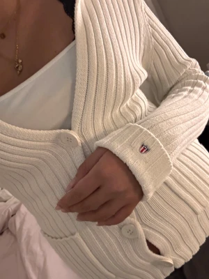 Vit ribbad kofta från GANT - Vit ribbad kofta från GANT med snygg v-ringning och fyra stora knappar framtill. Långärmad modell med två fickor och broderad logga på ärmslutet. Mjuk stickad bomull som sitter normal och har en clean, klassisk look.