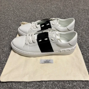Valentino Garavani Open sneakers vit/svart - Snygga Valentino Garavani Open sneakers i vitt skinn med bred svart läderdetalj över mitten. Klassisk rund tå, vita snören och ikoniska nitar på hälen. Kommer med original dustbag. Perfekta för dig som gillar stilrena och lyxiga sneakers. Dustbagen är fläckig vilket visas på bild 4 annars är det ett par riktigt fräscha skor🤩PRIS KAN DISKUTERAS!!! Säljer i befintligt skick, kan inte garantera att de är äkta då jag själv är andrahandsköpare.