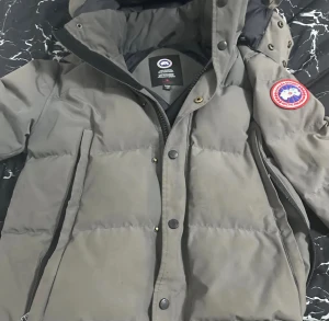 Grå dunjacka från Canada Goose - Säljer en grå dunjacka från Canada Goose med klassisk patch på ärmen. Jackan har huva med avtagbar pälskant, dragkedja och knappar framtill samt två stora fickor. Perfekt för kalla vinterdagar och riktigt snygg streetstil. Den är helt ny