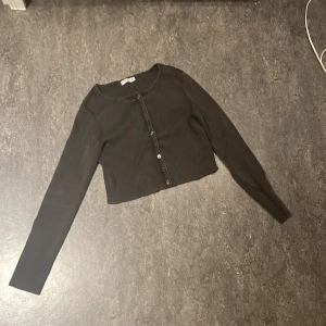 Svart croppad kofta från Stradivarius - Svart croppad kofta från Stradivarius i ribbad bomullsmix. Tröjan har lång ärm, rund hals och knappar framtill. Perfekt att styla med jeans eller kjol för en chill look. Enkel och stilren design som funkar året runt. Det är också en ”magtröja”.